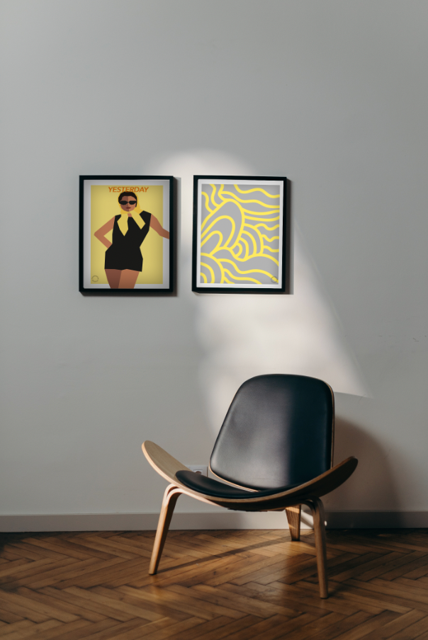 Billede af simpelt rum i med trægulv i en varm farve og en hvid væg. Der er en stol i træ og sort læder, og over hænger to Kreative Kunstplakater i grafisk design. Den ene af en kvinde med solbriller, den anden af et simpelt, abstrakt mønster.