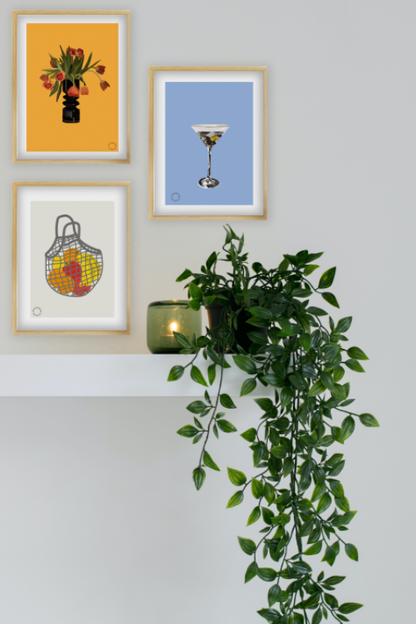 Billede af en hvid væg med en hvid hylde og en grøn plante på. Der hænger tre Kreative Kunstplakater i grafisk design. En plakat af en vase med tulipaner, en af et martiniglas og em af et net med frugter i