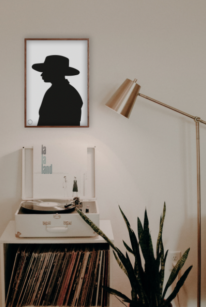 En hvid væg, hvor der står en pladespiller, en plante og en lampe. På væggen hænger der en Kreativ Kunstplakat i grafisk design af omridset af en mand med hat i profil